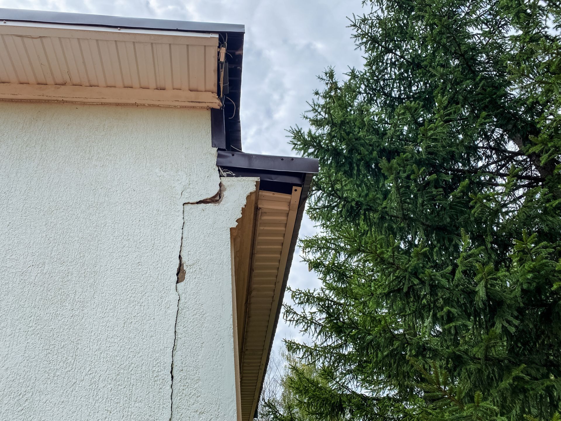 Cracked stucco exterior wall under a roof eave beside a tall evergreen tree