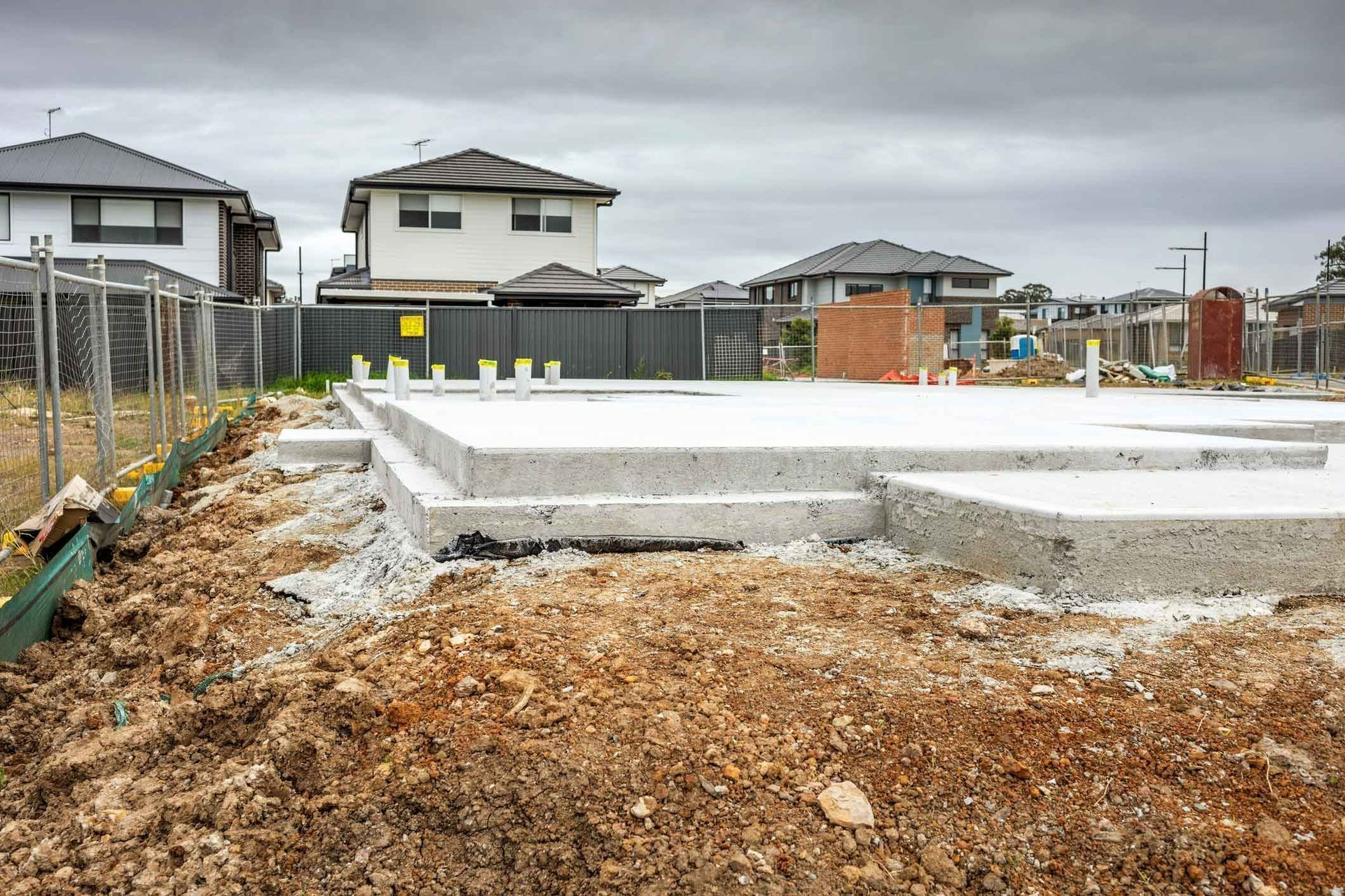 Foundation slab under construction in a new housing development, with dirt and fenced lots on an overcast day