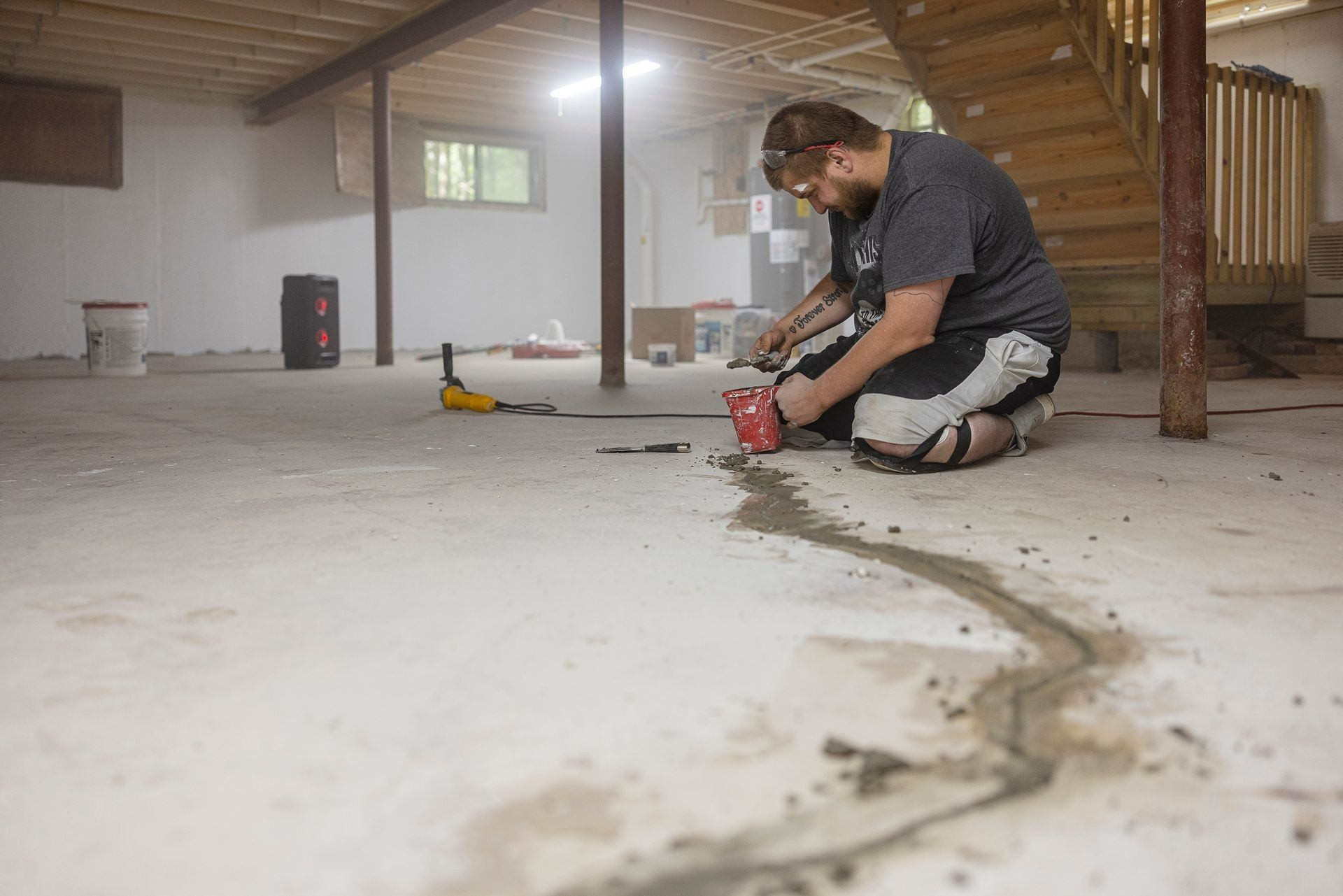 Worker kneeling on a basement floor, sealing a long crack with red caulk under wooden stairs.