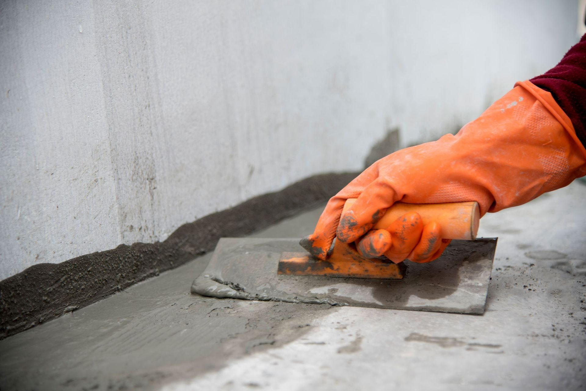 Orange-gloved hands trowel wet concrete along a wall indoors