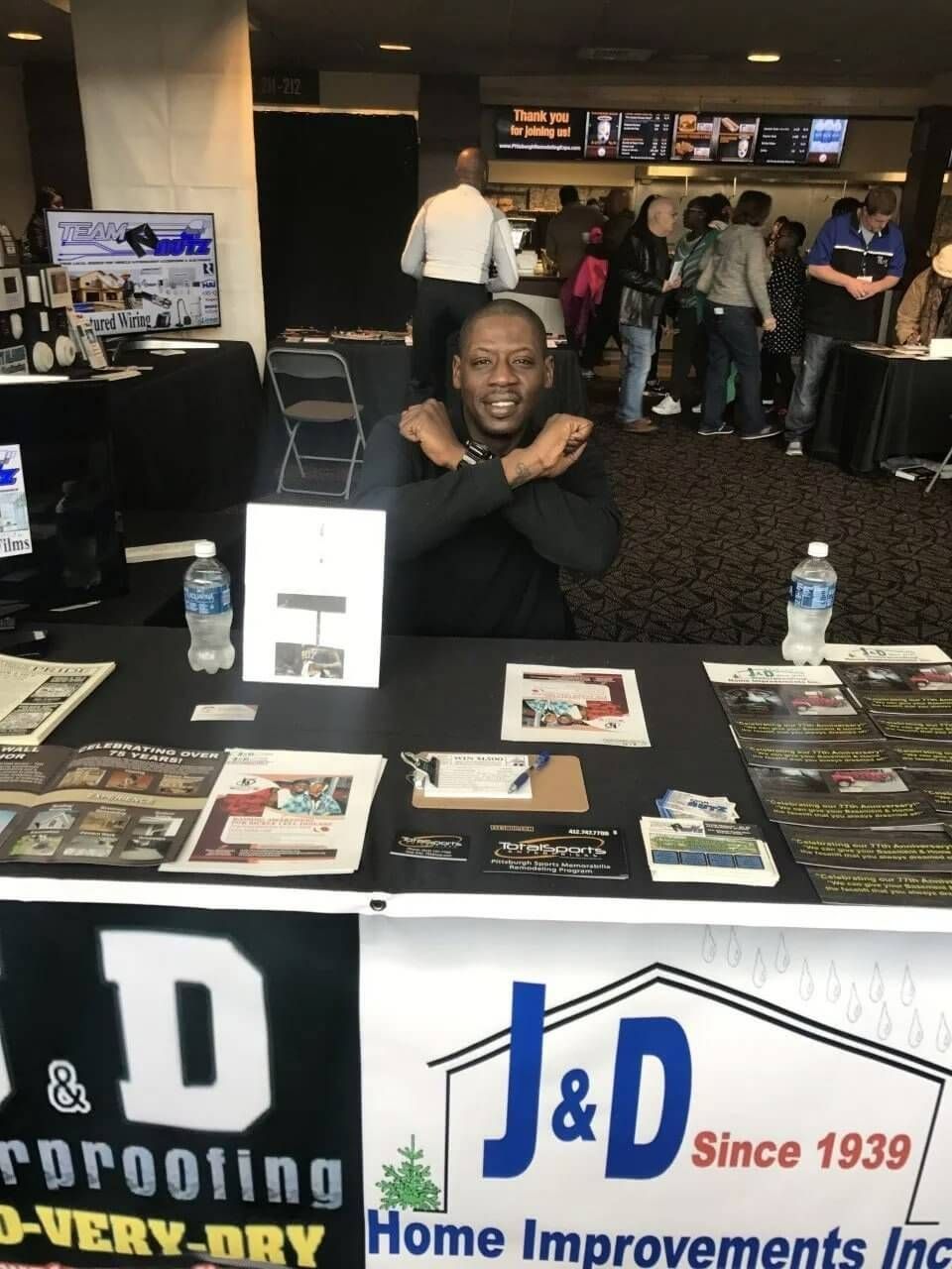 Man smiling at a trade show booth with J&D Waterproofing signage and brochures on display