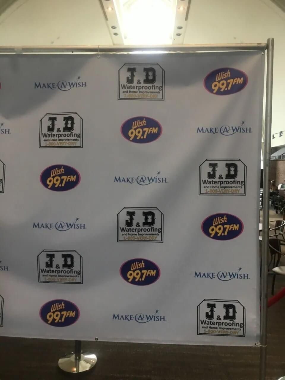 Step-and-repeat banner with J.D. and 99.7 FM logos on a white backdrop, standing on a floor stand