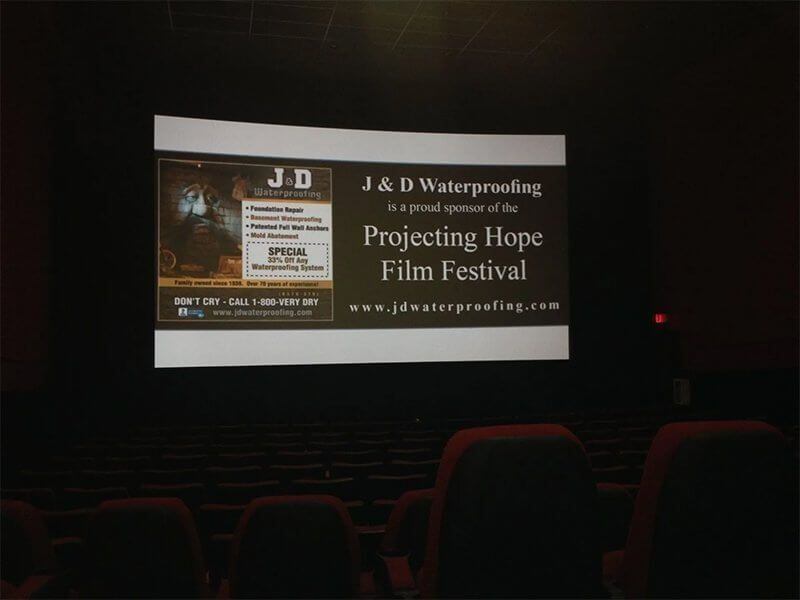 Movie theater screen showing an ad for the J&D Waterproofing Projecting Hope Film Festival.