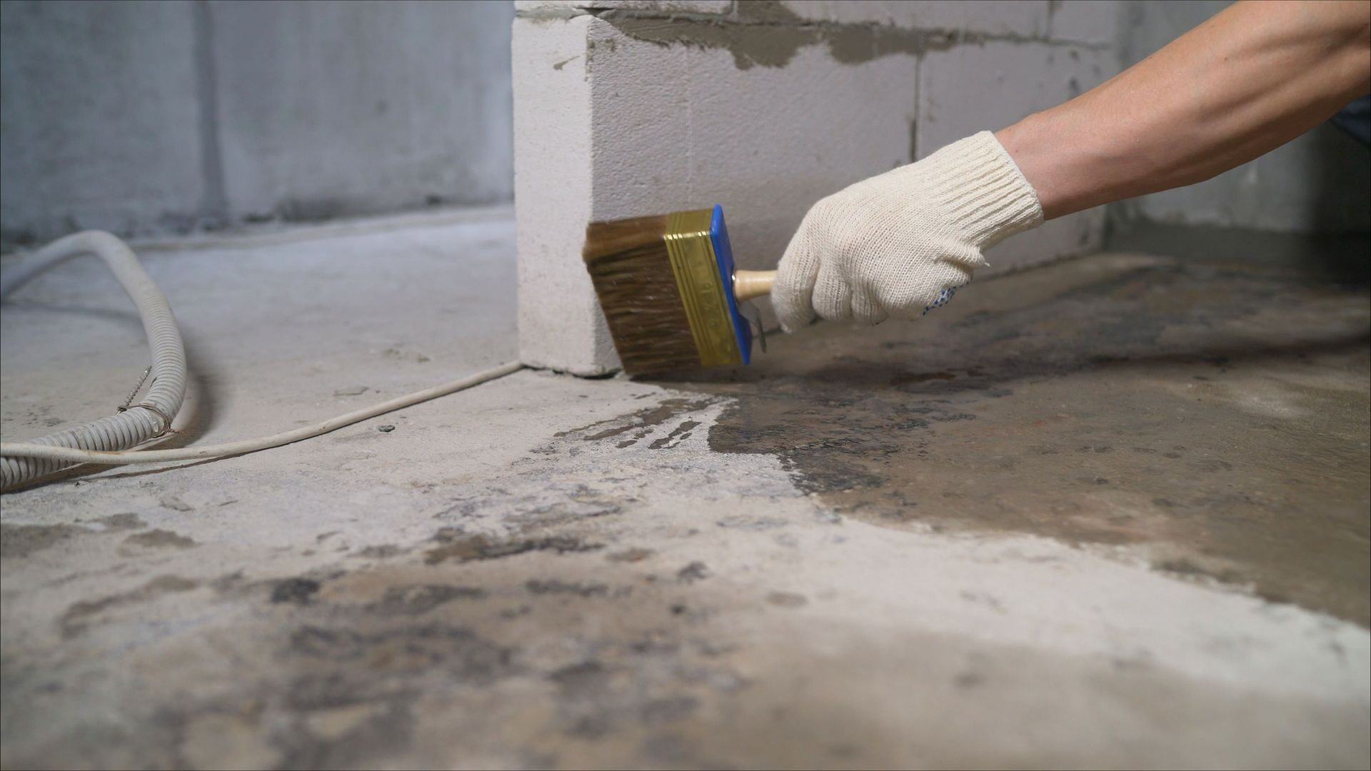 Gloved hand using a brush to apply sealant along a concrete floor and wall seam