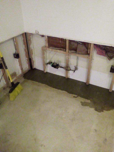 Partially demolished basement wall with exposed studs, pipes, and tools on a concrete floor