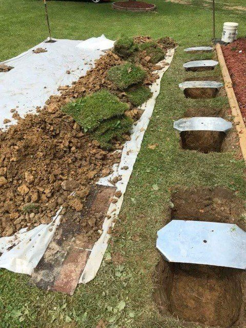 Construction trench with white tarp, exposed soil, and concrete footing holes along a grassy lawn