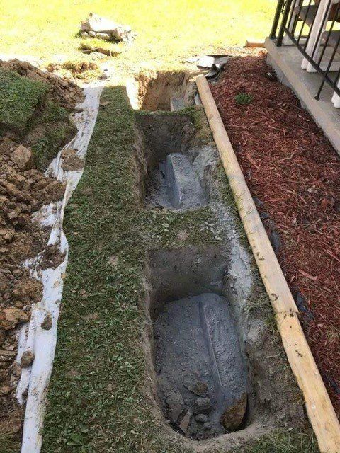 Four narrow, dug-out trenches lined with grass and concrete in a yard, beside a wooden border and soil bed.