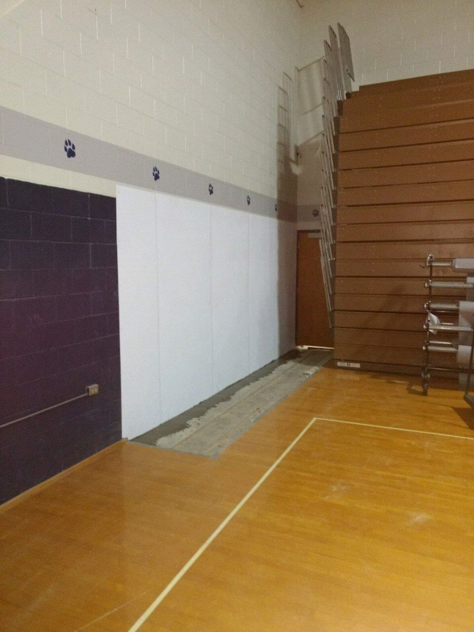 Empty gymnasium with wooden floor, wall padding, and stacked bleachers in the corner