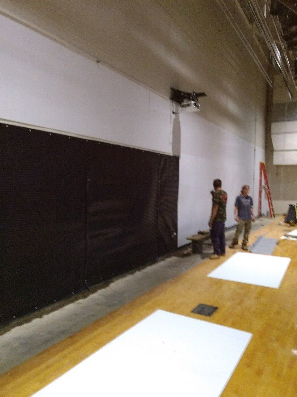 People working in a large gymnasium with rolled white floor mats and a red ladder against the wall.
