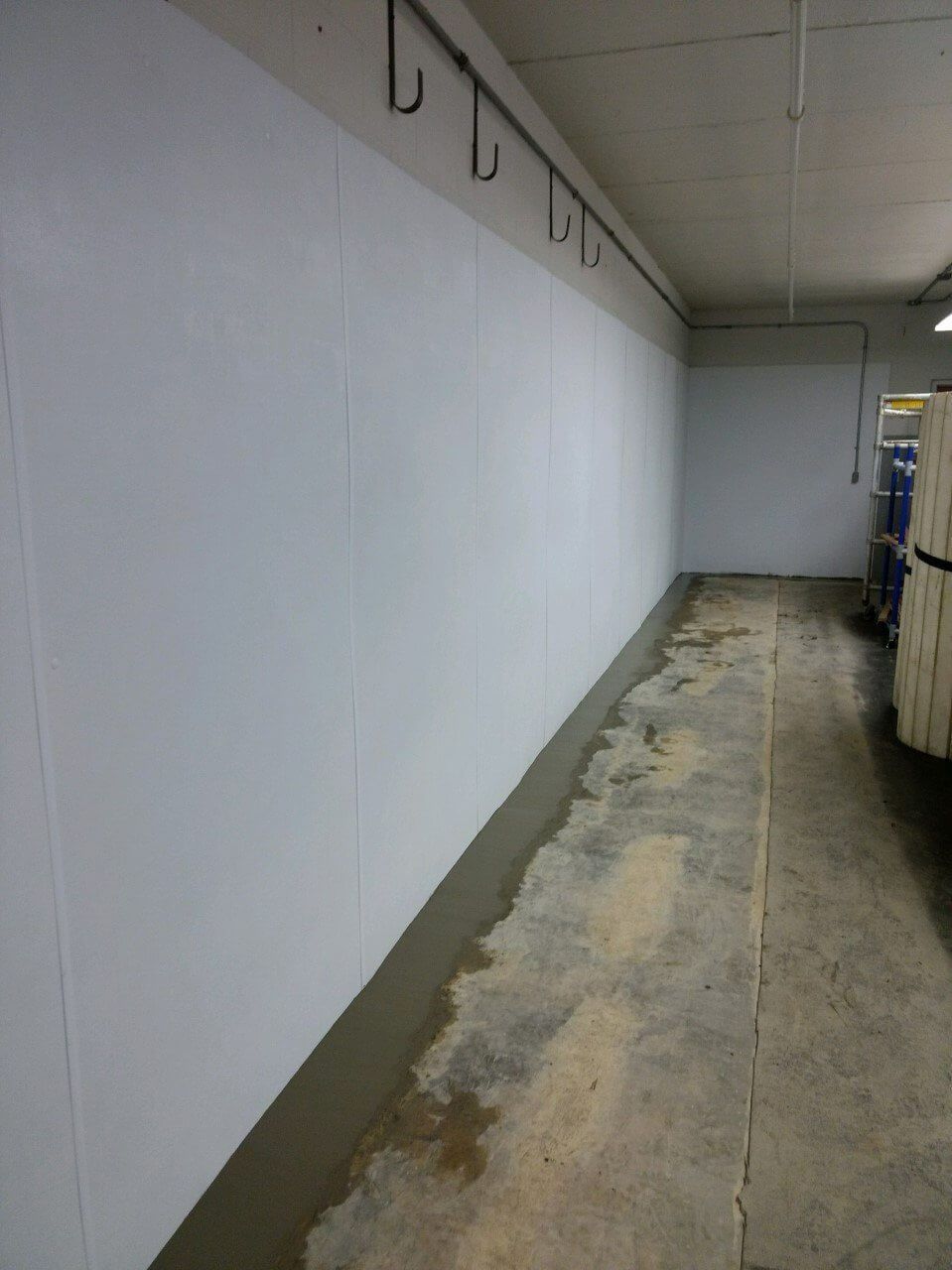 Narrow hallway with white walls, hooks, and a worn, stained floor in a utilitarian interior.