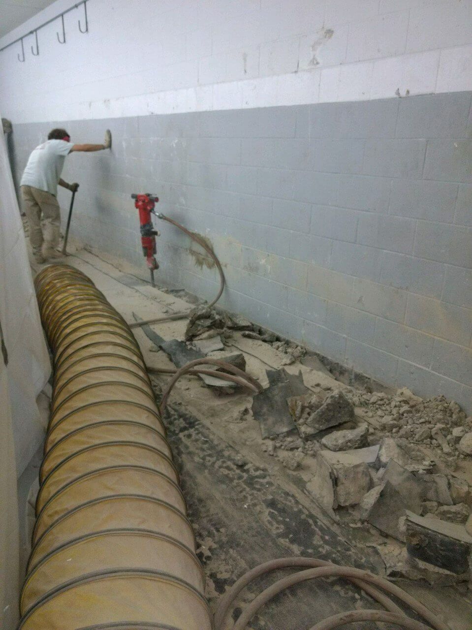 Worker in tunnel beside a large yellow hose and red pipe, clearing rubble along a wall.