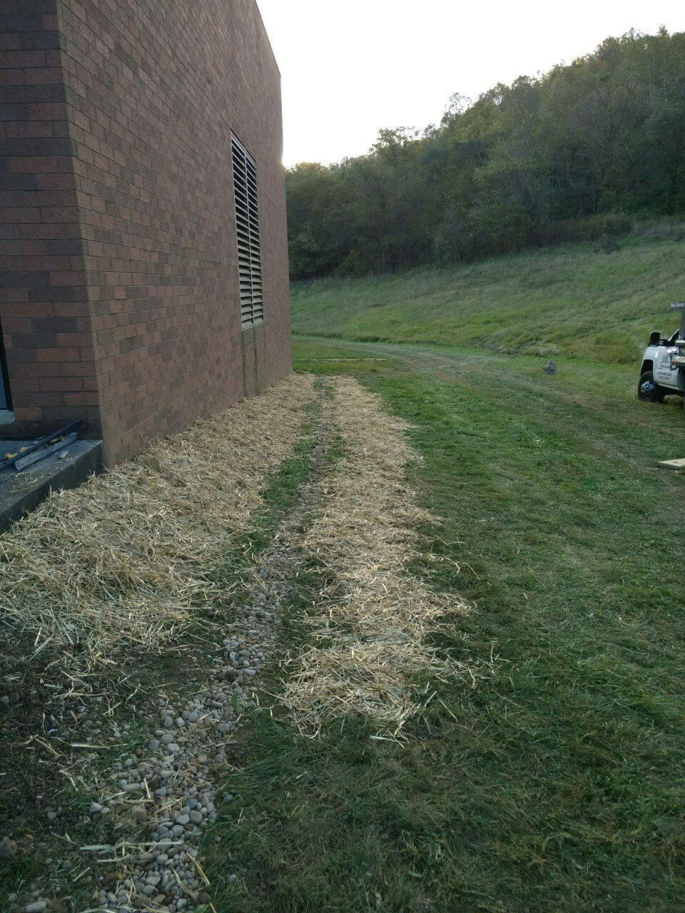 Brick building beside grassy slope and hill, with a small white vehicle on the right.