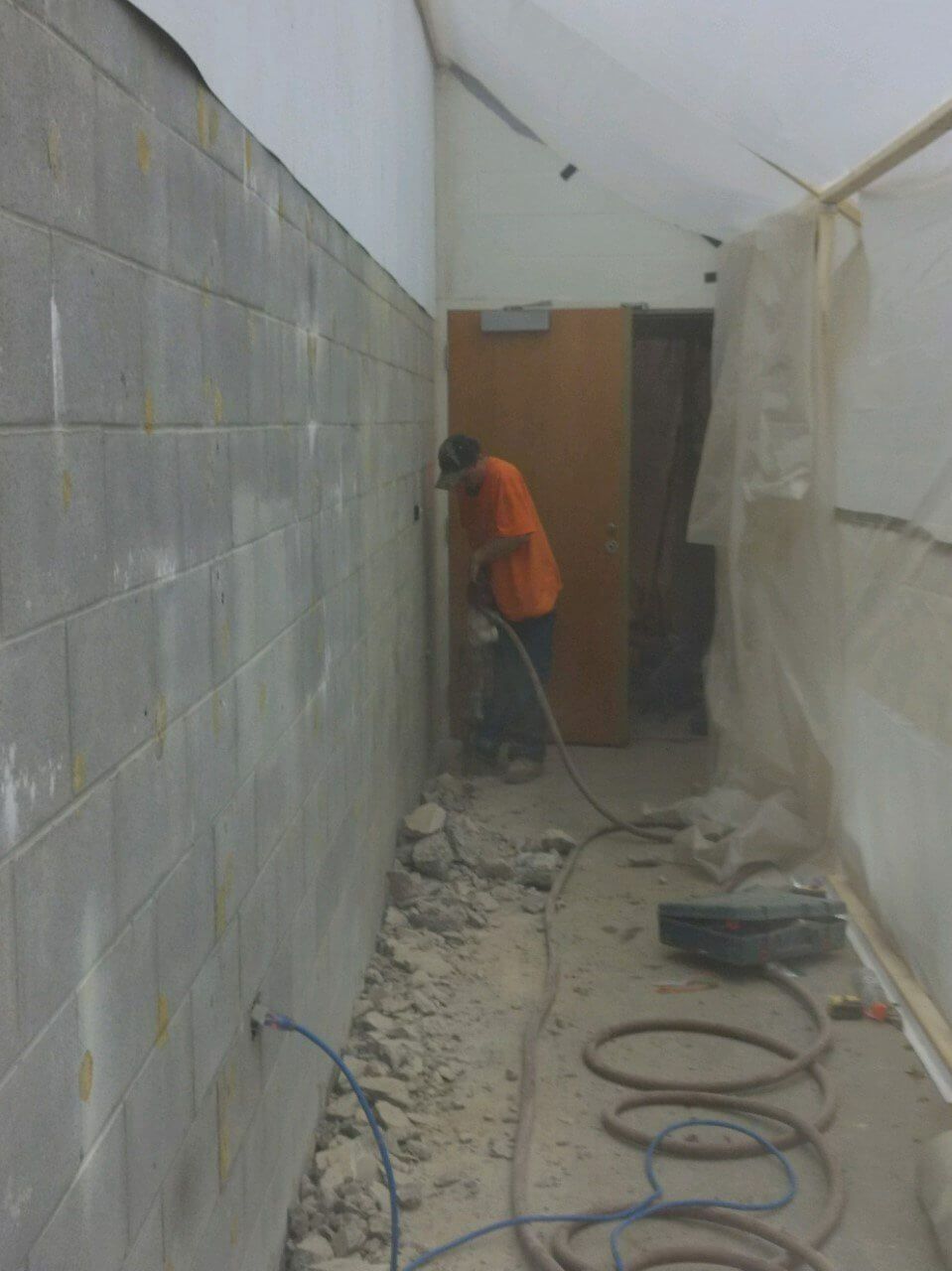 Worker using a jackhammer to break concrete in a narrow hallway with dust and debris