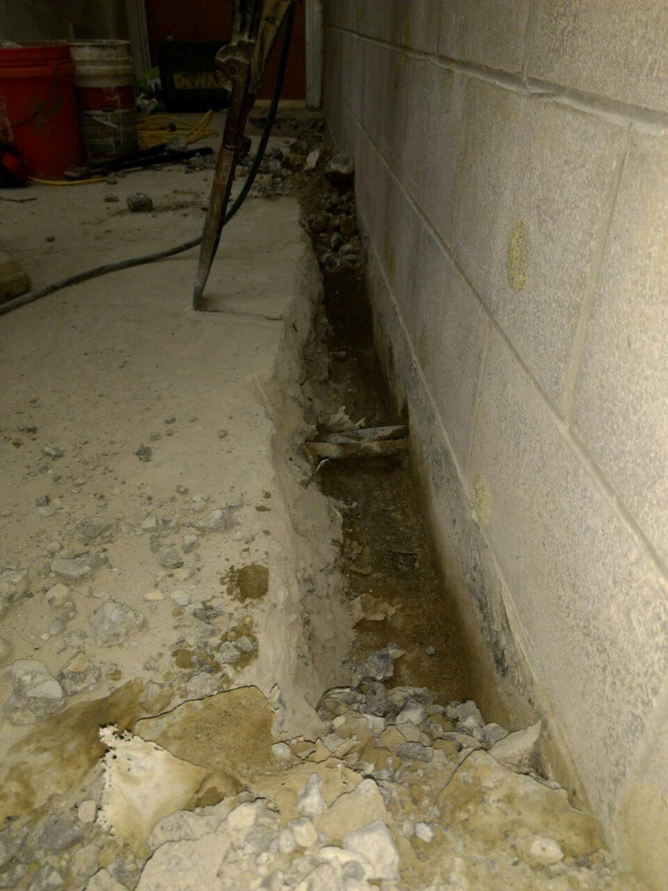 Cracked basement floor beside cinderblock wall with debris and exposed soil along the foundation