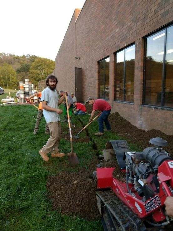 People digging a trench beside a brick building, with tools and a red machine on grass.