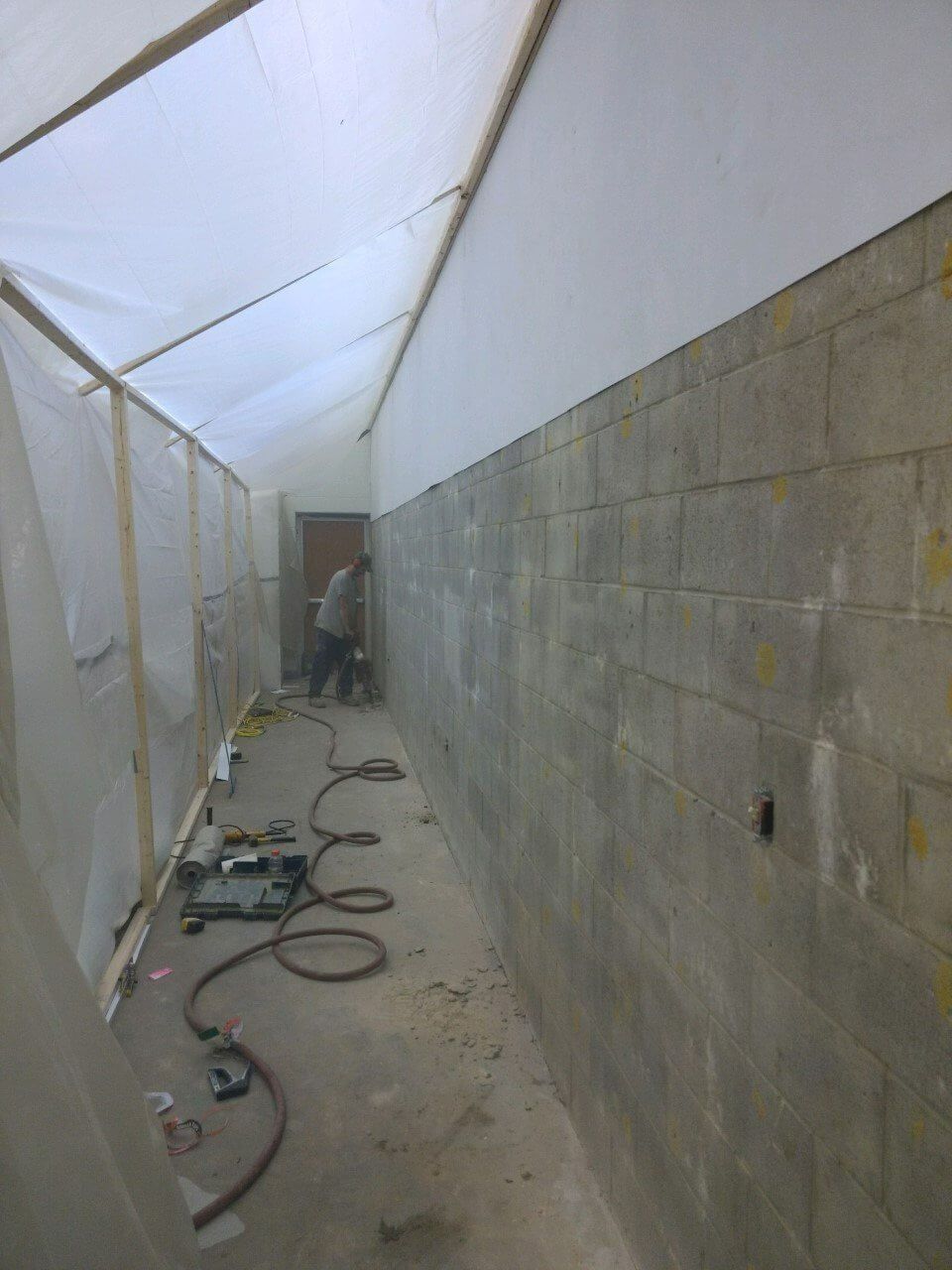 Narrow unfinished corridor with cinderblock walls, white tarps overhead, and hoses scattered on the floor.