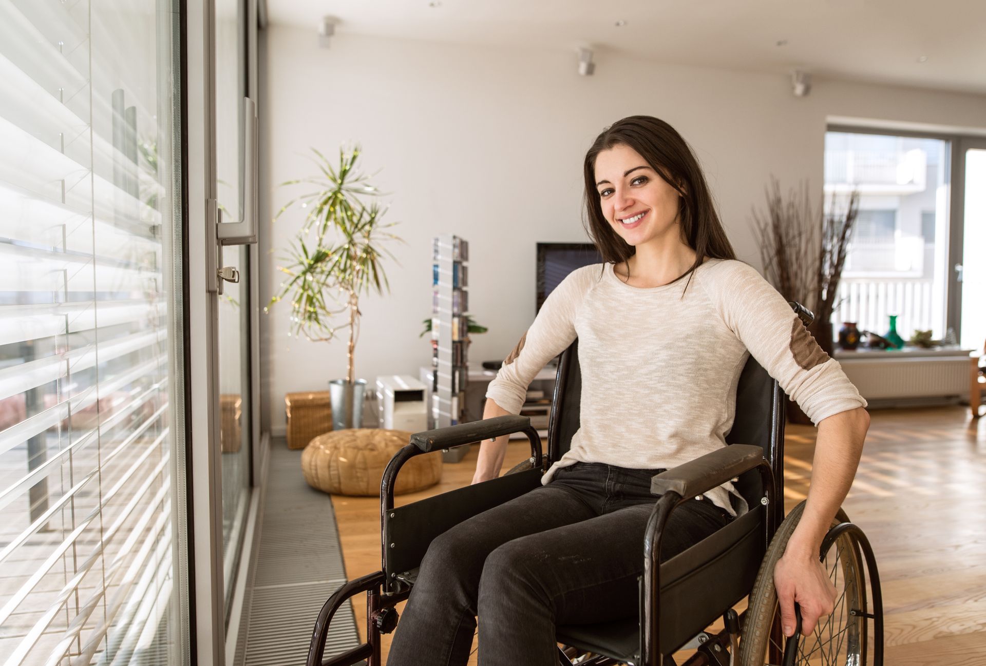 Woman in a wheelchair smiles at the camera indoors near a window; bright room with a plant.