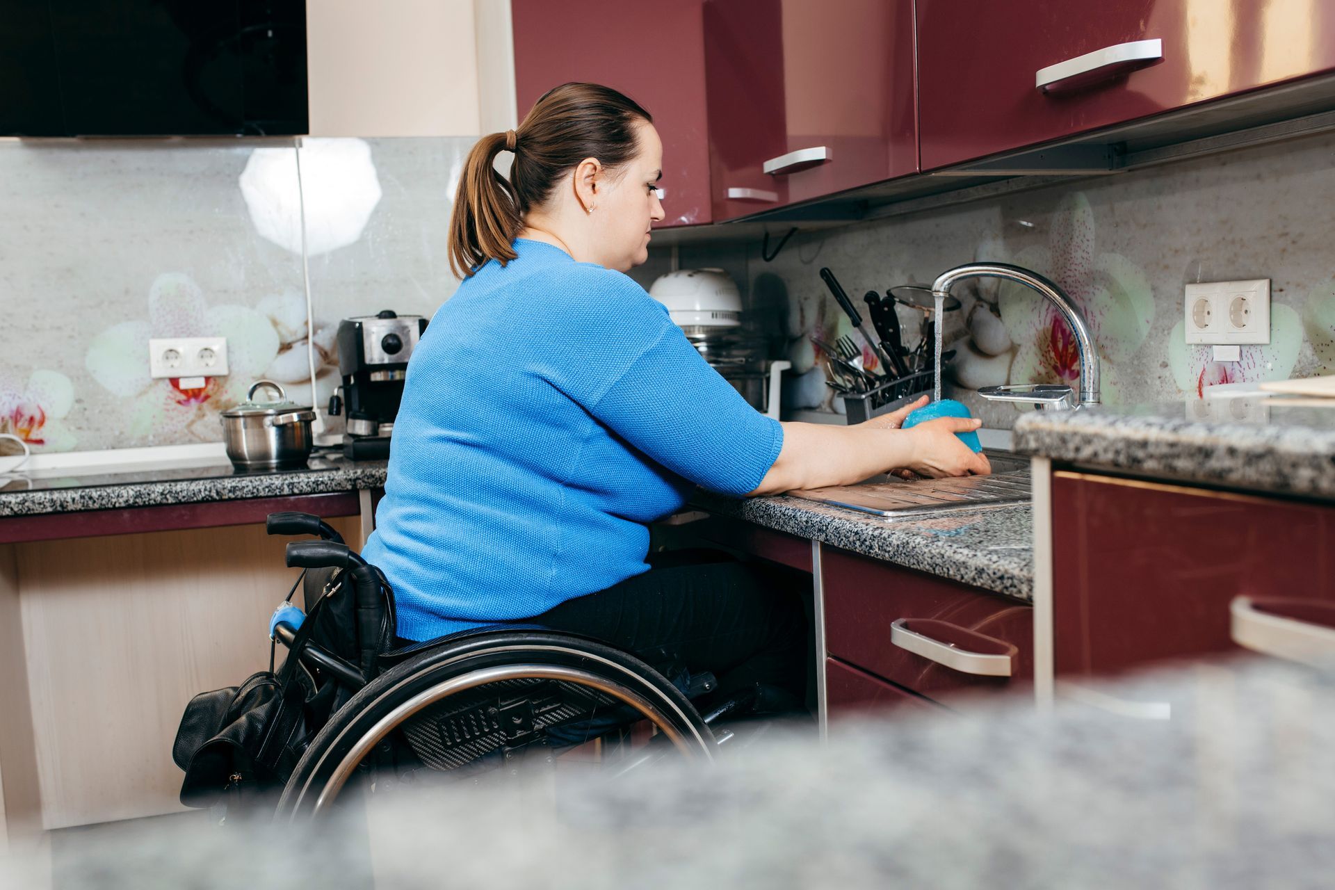A woman in a wheelchair is washing dishes in the kitchen.