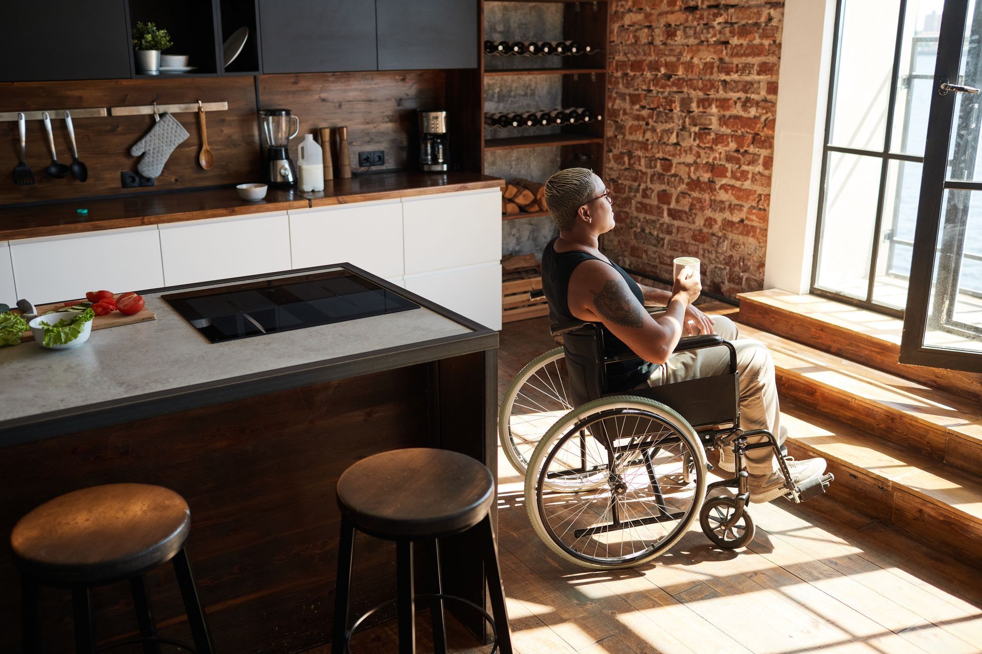 A woman in a wheelchair is sitting in a kitchen drinking a cup of coffee.