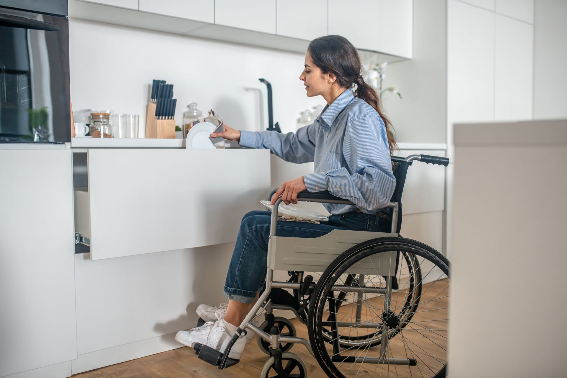 A woman in a wheelchair is reaching into a drawer in a kitchen.