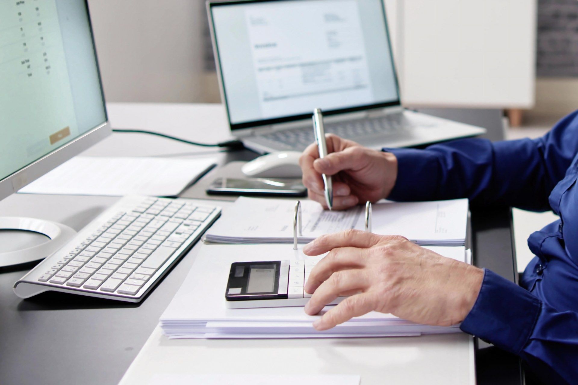 Person in blue shirt using a calculator, looking at documents with computer and laptop in office setting.