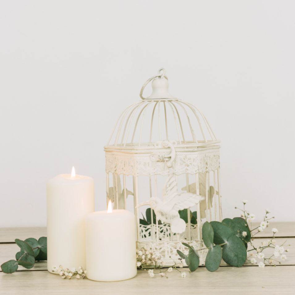 Dos velas blancas encendidas y una jaula de pájaros adornada sobre una superficie de madera, con vegetación y flores blancas.