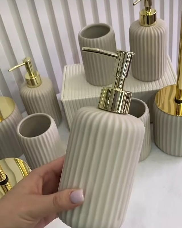 Mano sosteniendo un dispensador de jabón acanalado de color beige con bomba dorada, rodeado de accesorios de baño a juego.