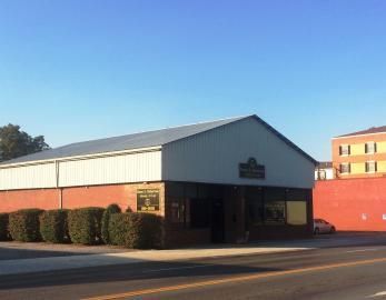 A brick and metal-sided commercial building with a row of small shrubs, viewed from a street corner under a blue sky.