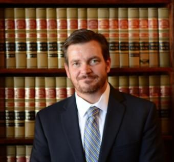 A person in a suit and tie smiling in front of a wooden bookshelf filled with law books.