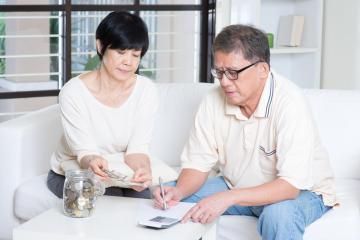Two people sit on a white sofa, reviewing financial documents and counting coins from a jar to manage their budget.