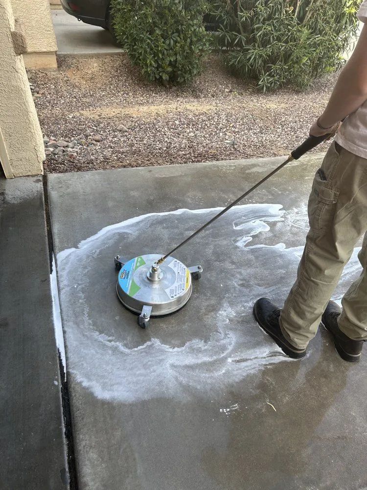 Person pressure washing a concrete surface with a rotating cleaner, creating foamy suds outdoors.