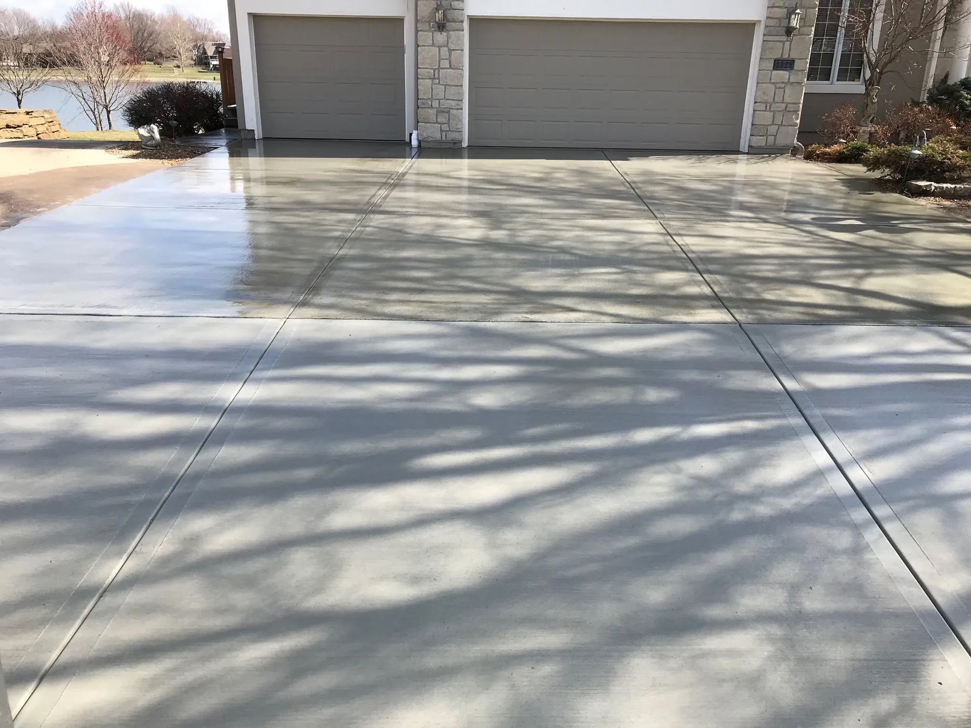 A gray concrete driveway leading to a house with two garage doors; sunlight casts shadows.