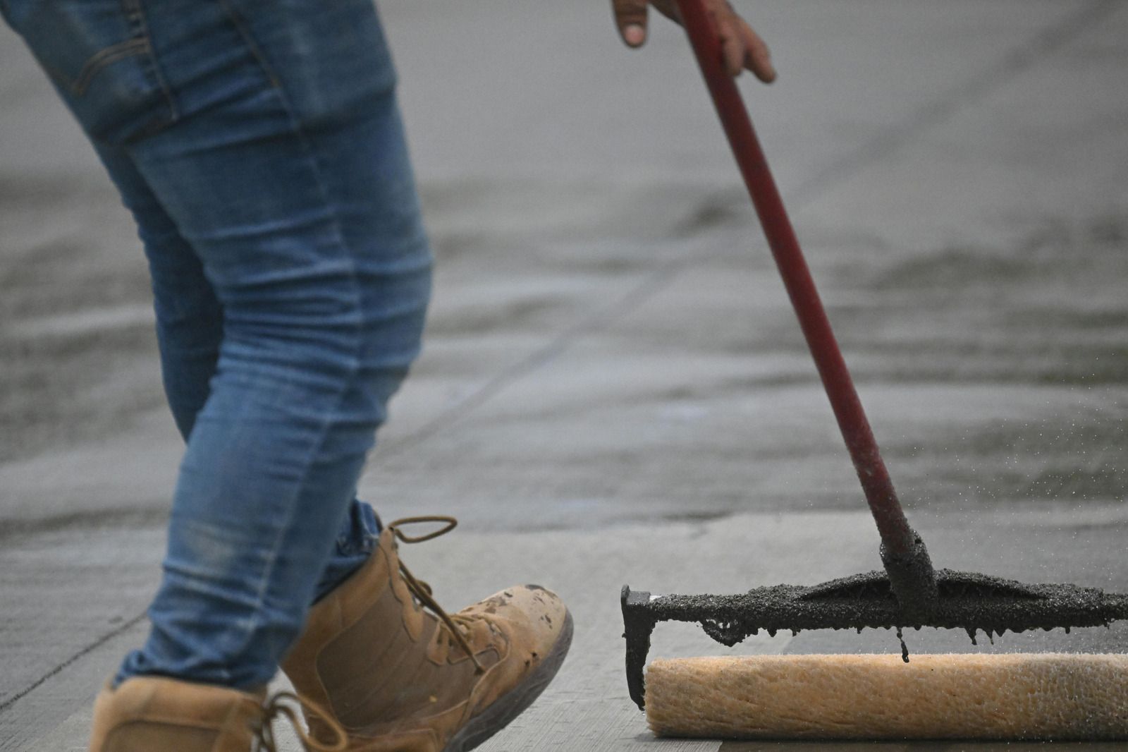 Person in jeans and work boots rolling asphalt on a flat roof with a long-handled roller.