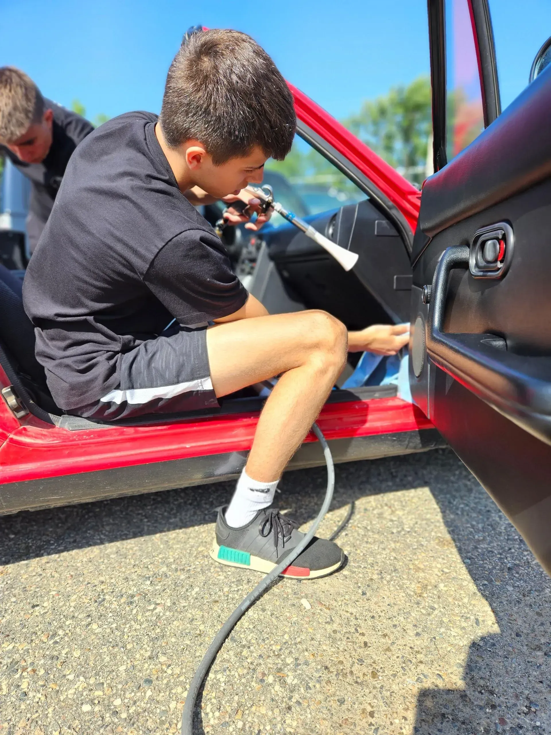 A young man is sitting in the door of a red car using a hose.