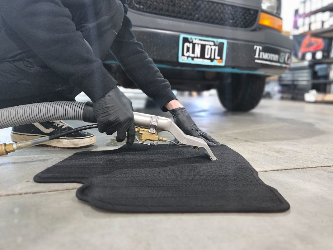 Person wearing black gloves vacuums a black car floor mat on a concrete floor near a black vehicle.