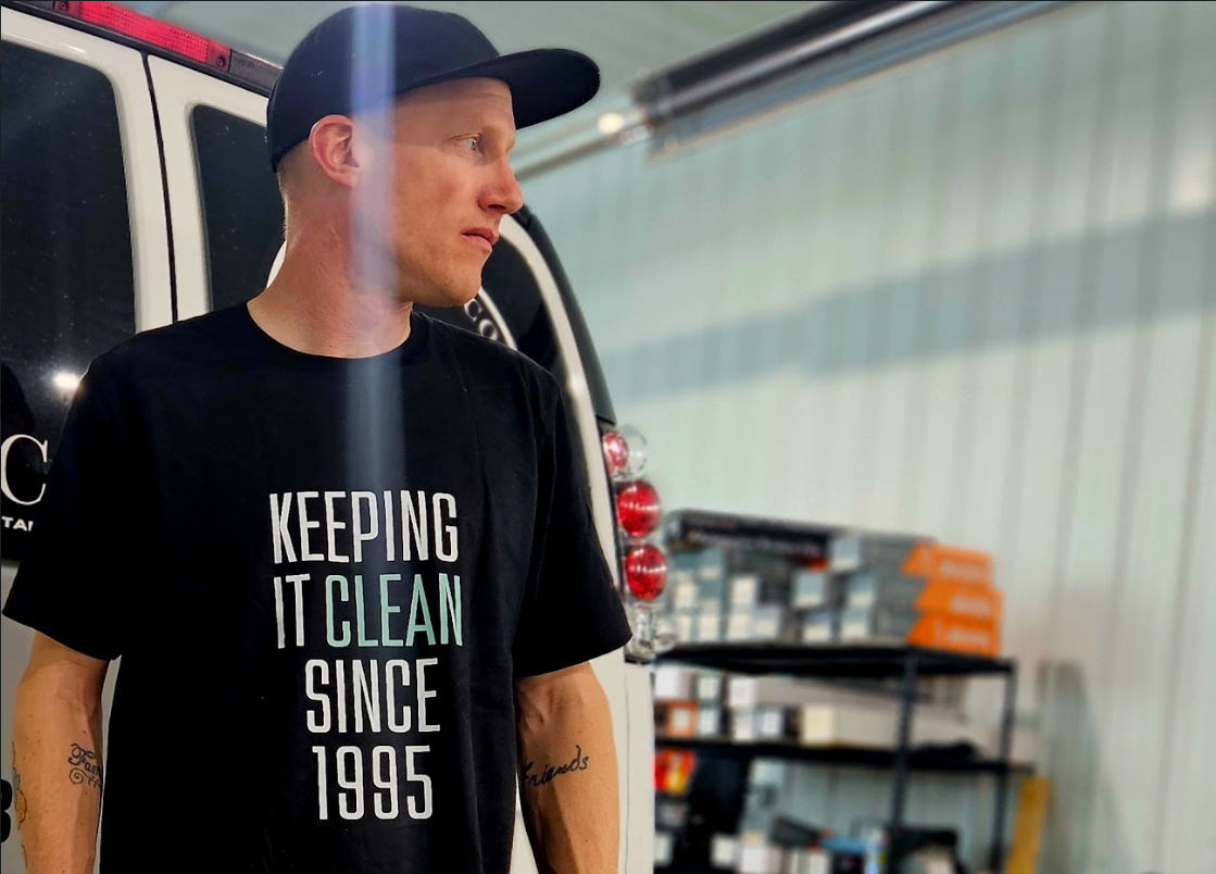 A man wearing a black t-shirt and cap poses in a garage. His shirt reads