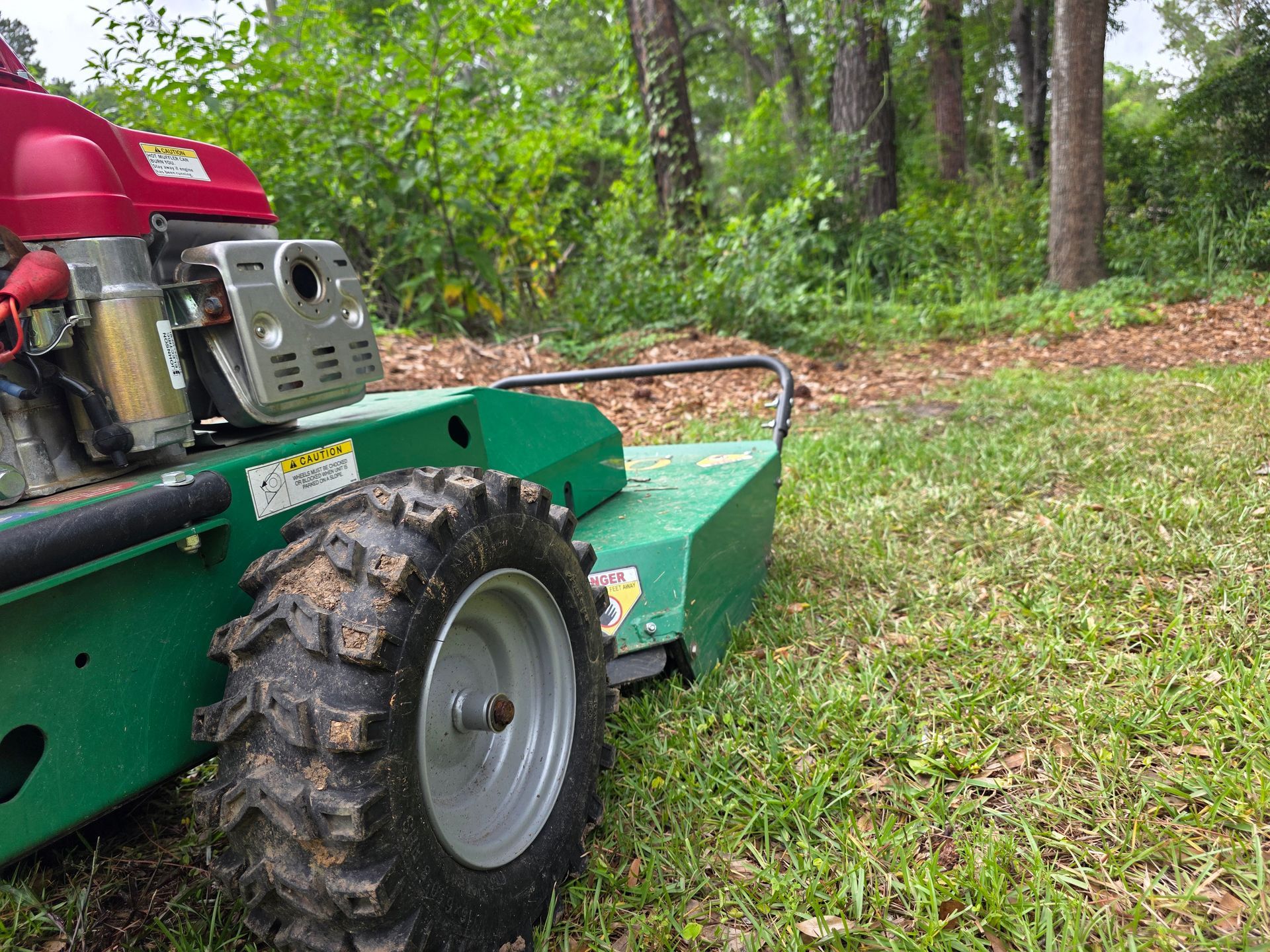Green brush mower with a red engine in a grassy area with trees.