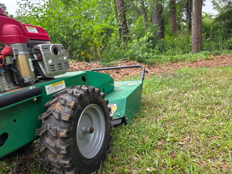 Green brush mower with a red engine in a grassy area with trees.