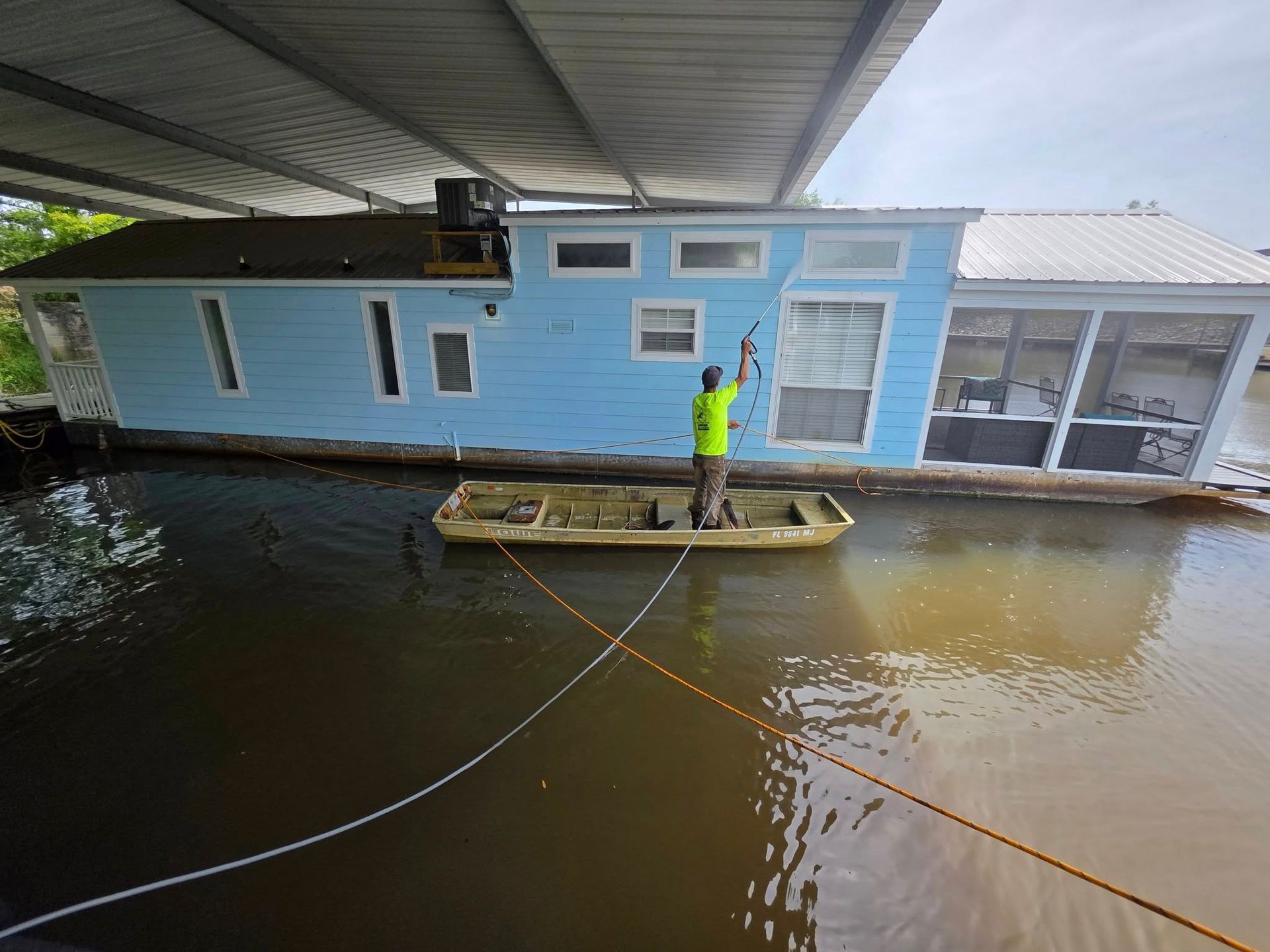 A man in a boat near a flooded blue houseboat under a bridge, using a pole.