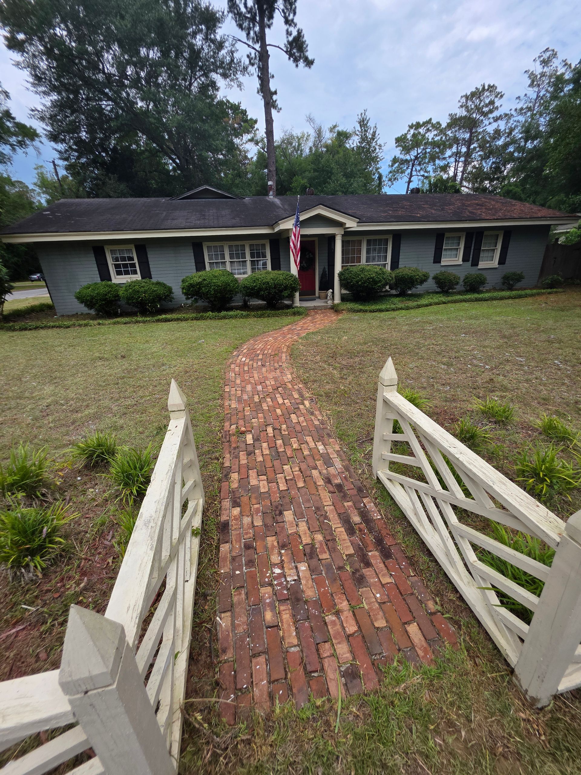 Brick path leads to a blue house with black shutters, white picket fence, and overgrown lawn.
