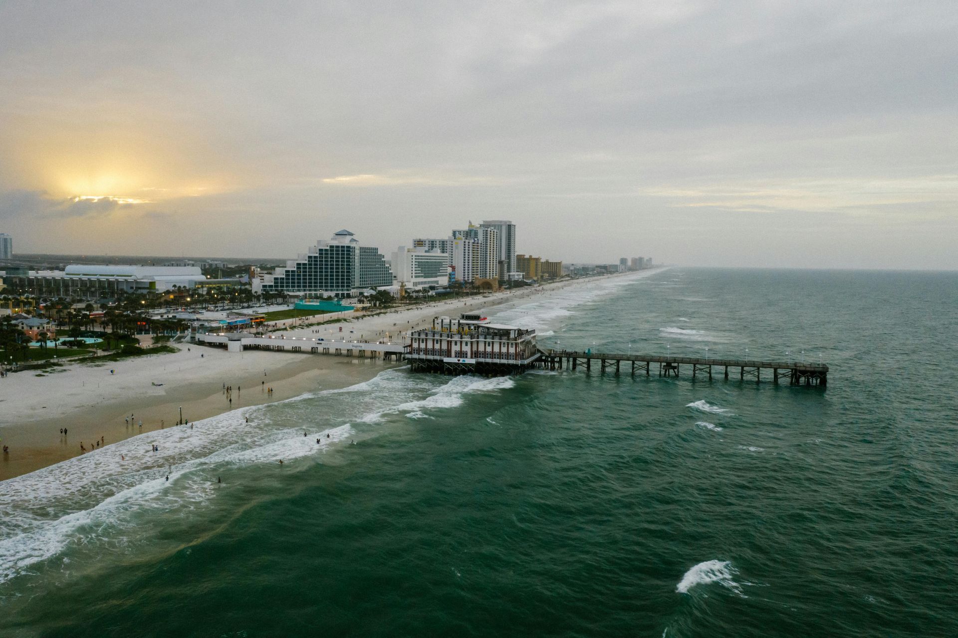 Beach scene with pier, waves, buildings, and cloudy sky.