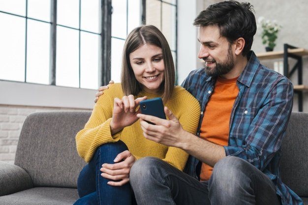 Una pareja está sentada en un sofá gris, sonriendo mientras miran juntos un teléfono inteligente 