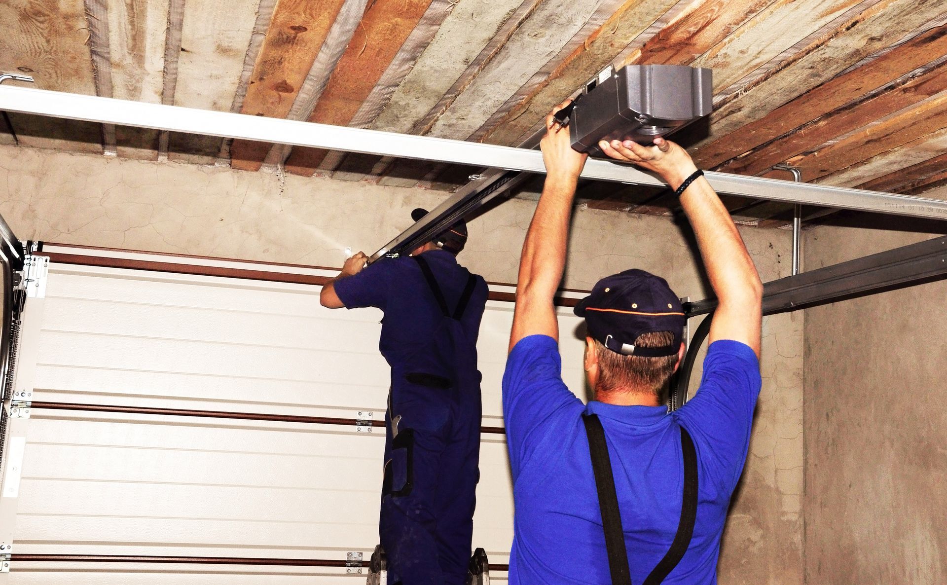 Two workers installing a ceiling track in a garage, one in blue overalls and one on a ladder.