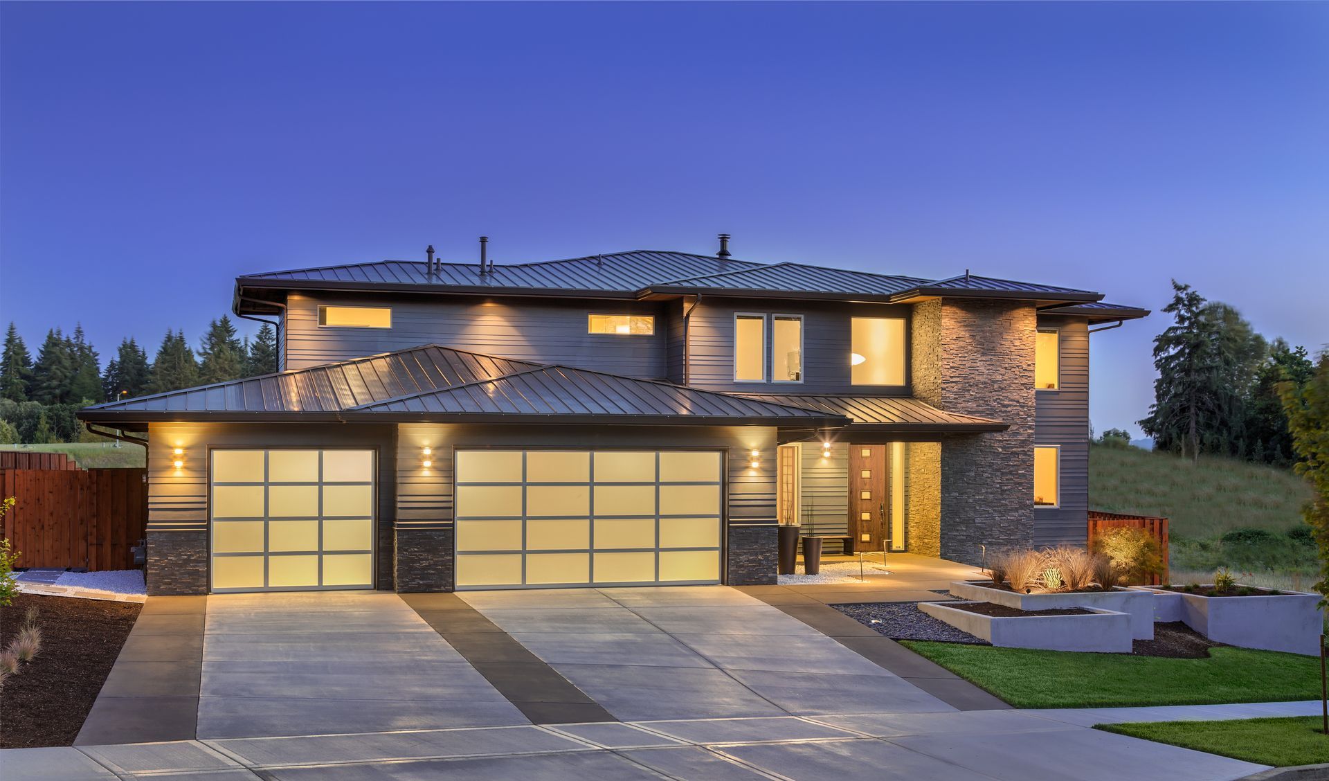 Modern two-story stone house at dusk with illuminated windows and a three-car garage