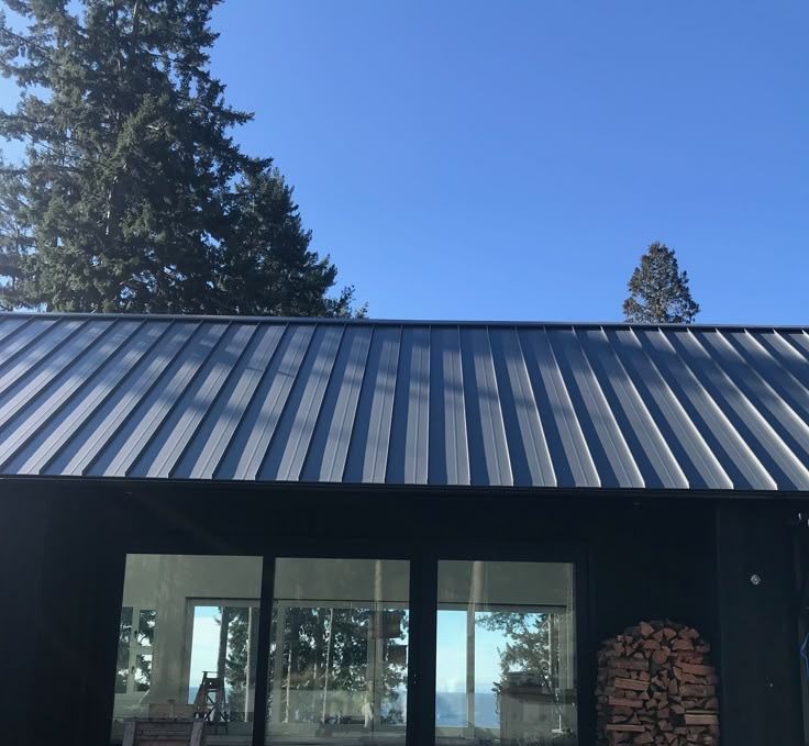 Modern black home with vertical standing seam metal roofing, large glass doors, and a minimalist concrete stepping stone path over gravel. Firewood is neatly stacked beside the entrance. — Apex Roofing Group in Byron Bay, NSW