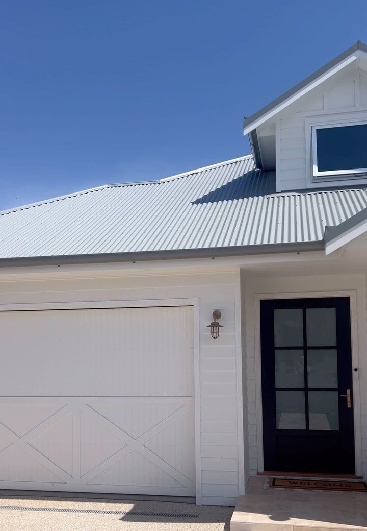 Contemporary home with light grey corrugated metal roofing, skylight feature, and soft blue weatherboard cladding paired with a white garage door and trim.— Apex Roofing Group in Ballina, NSW