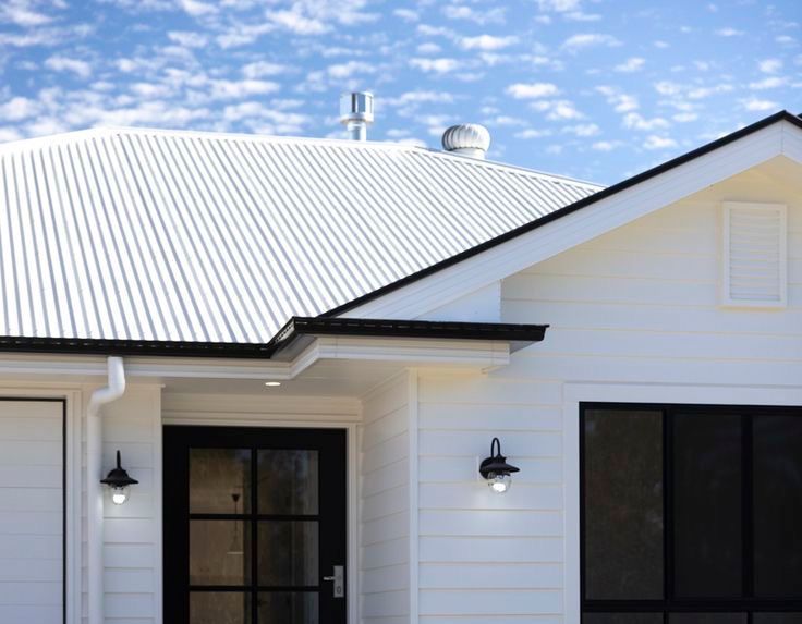 Front exterior of a modern white house with a corrugated metal roof, black front door, decorative block mailbox wall, and neatly landscaped yard under a blue sky. — Apex Roofing Group in Byron Bay, NSW