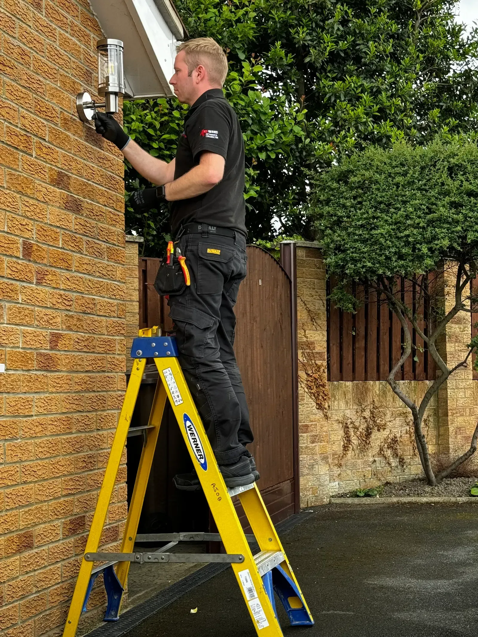 An AJOB Electrical Ltd electrician is standing on a ladder working on an outside light.