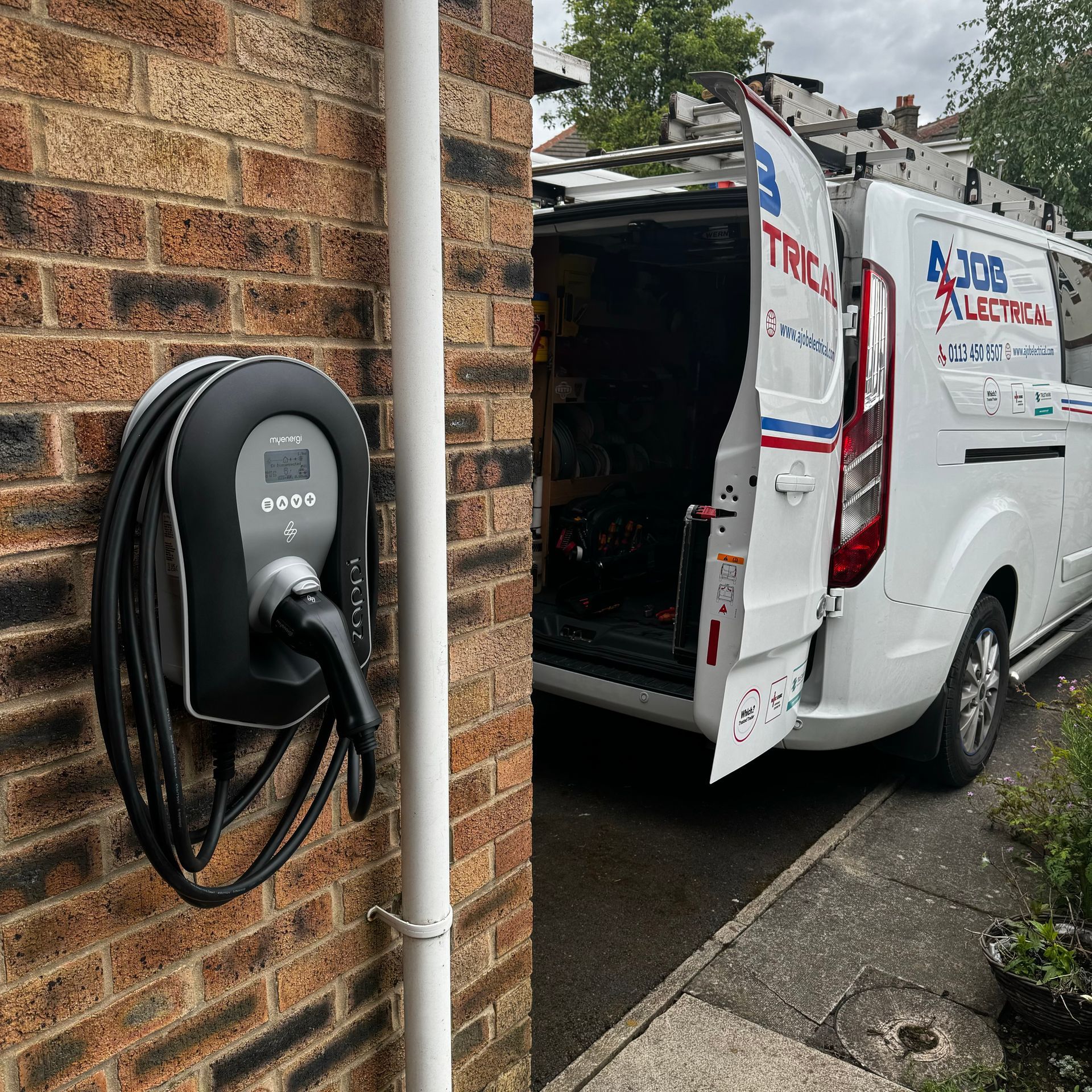 A white van is parked next to a brick wall with a charging station attached to it.
