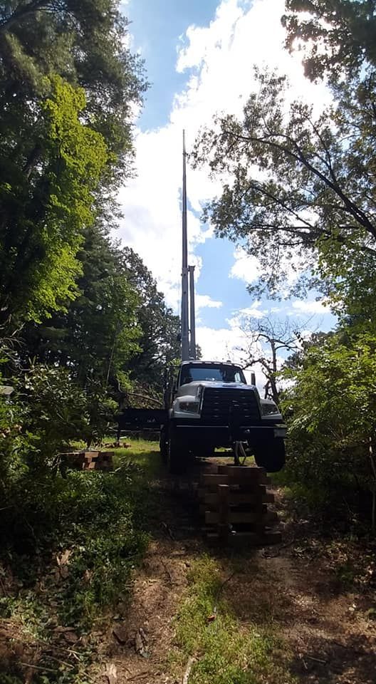 A large black truck on a dirt road, beneath a tall tower surrounded by trees. Blue sky.